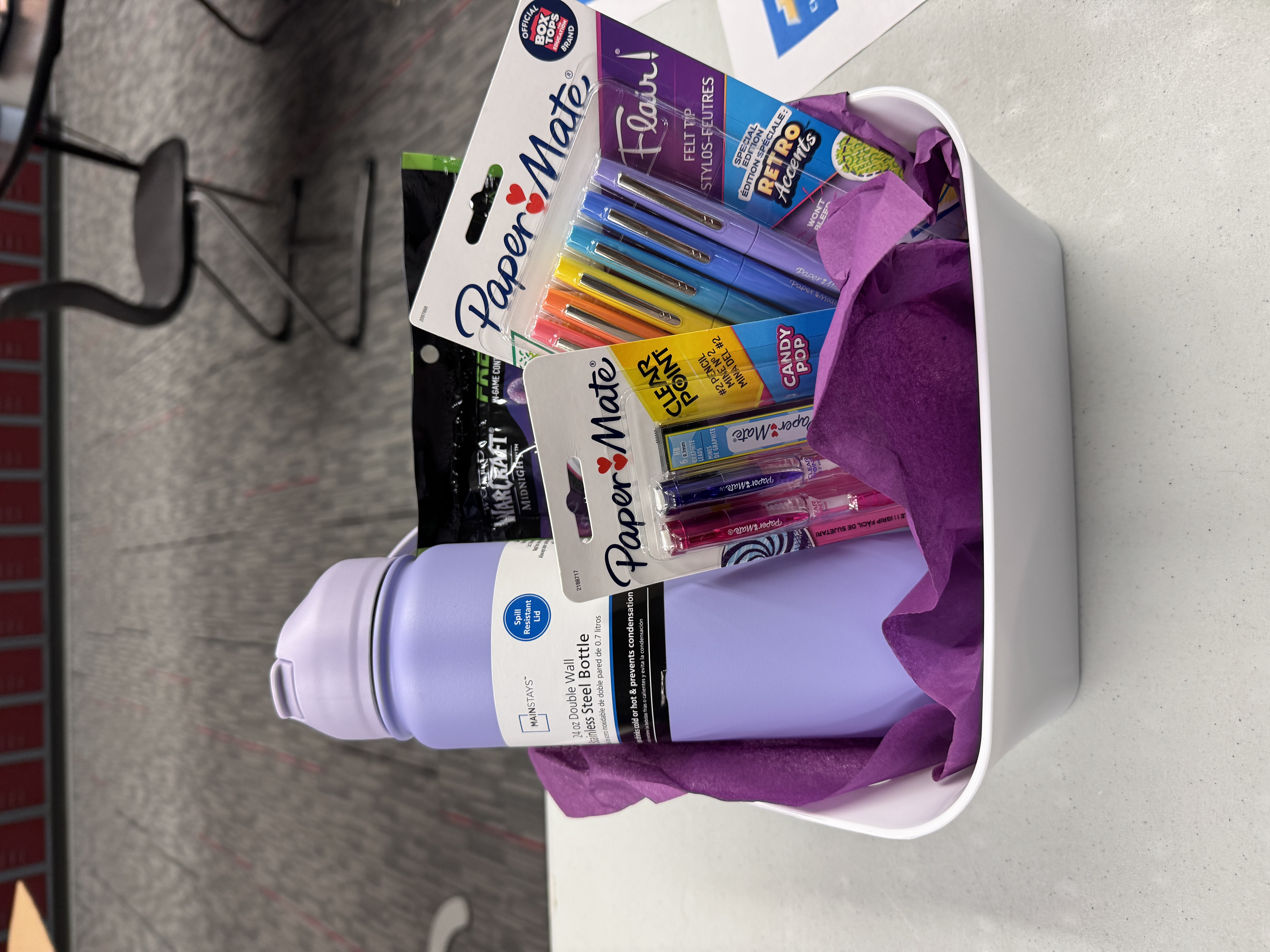 A white basket filled with colorful Paper Mate pens, a purple water bottle, and other stationery items.