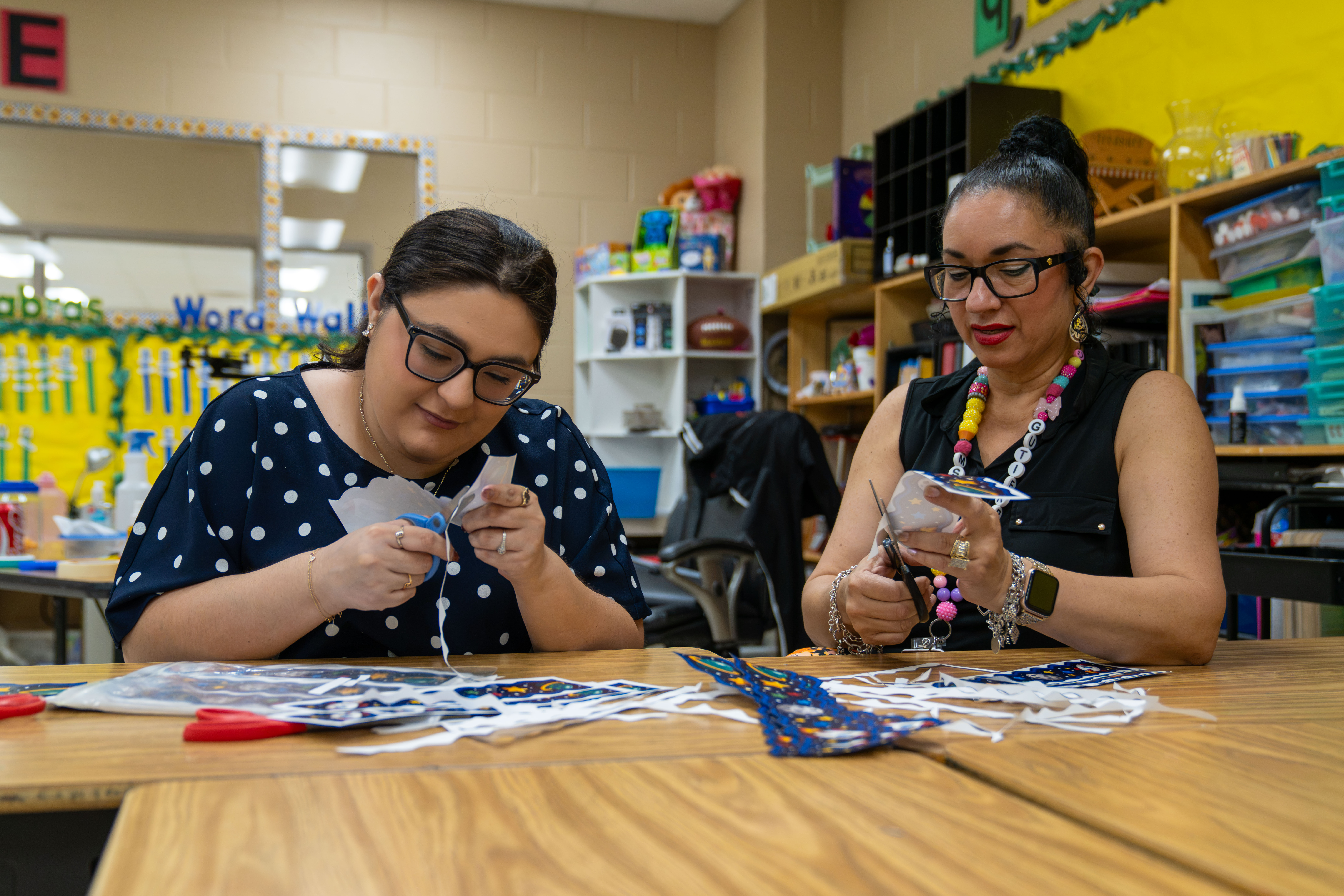 Two women are cutting out paper crafts at a wooden table in a classroom.