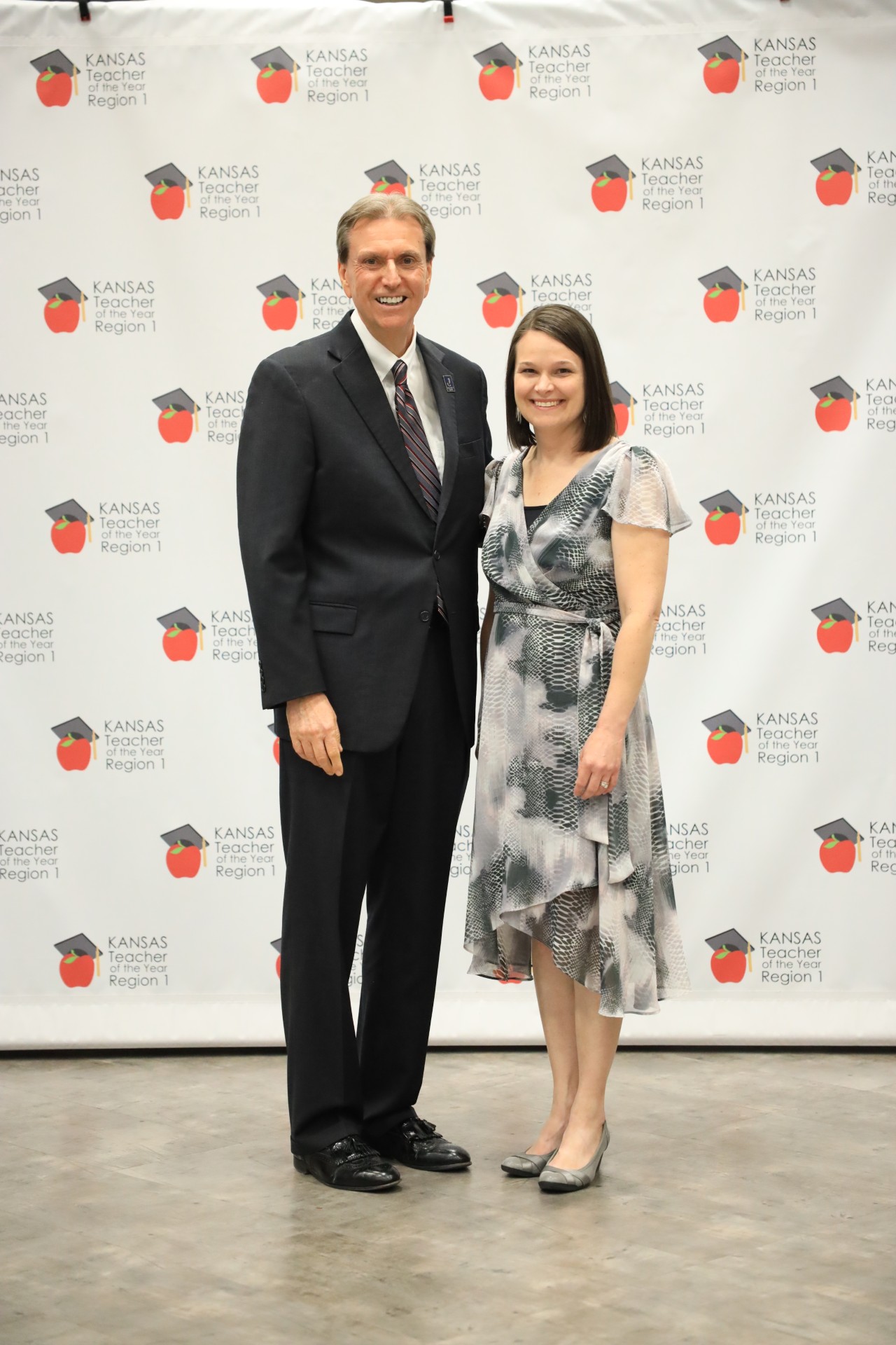A man in a suit and a woman in a patterned dress stand smiling in front of a backdrop with "Kansas Teacher of the Year Region 1" repeated.