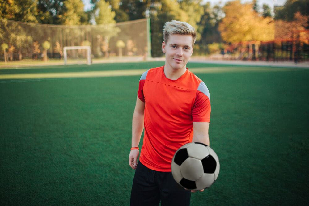 A young man with blonde hair holds a soccer ball on a green field.