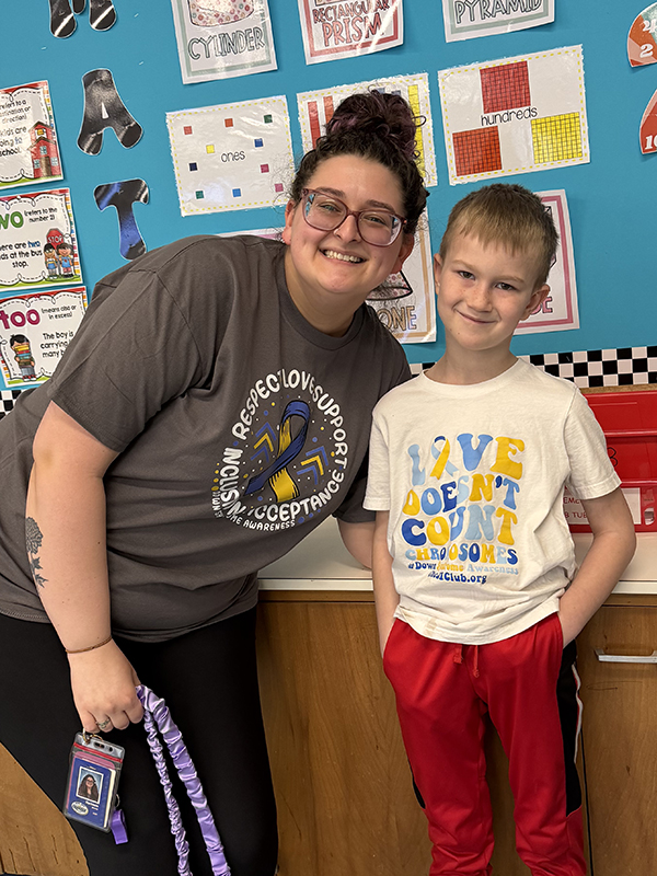 A smiling woman and a young boy stand together in a classroom with educational posters on the wall.