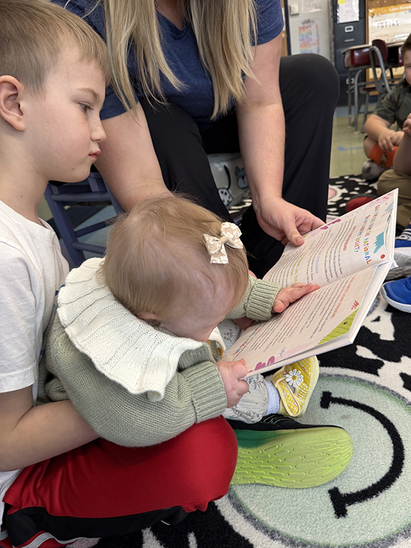 A group of children and an adult sit on a colorful rug, listening to a story from an open book.
