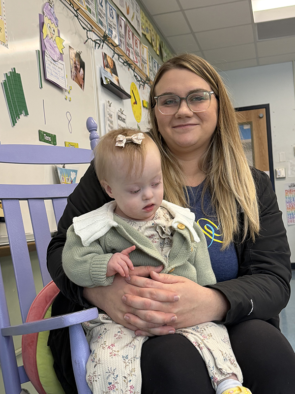 A woman holds a baby on her lap while sitting in a purple rocking chair in a classroom.