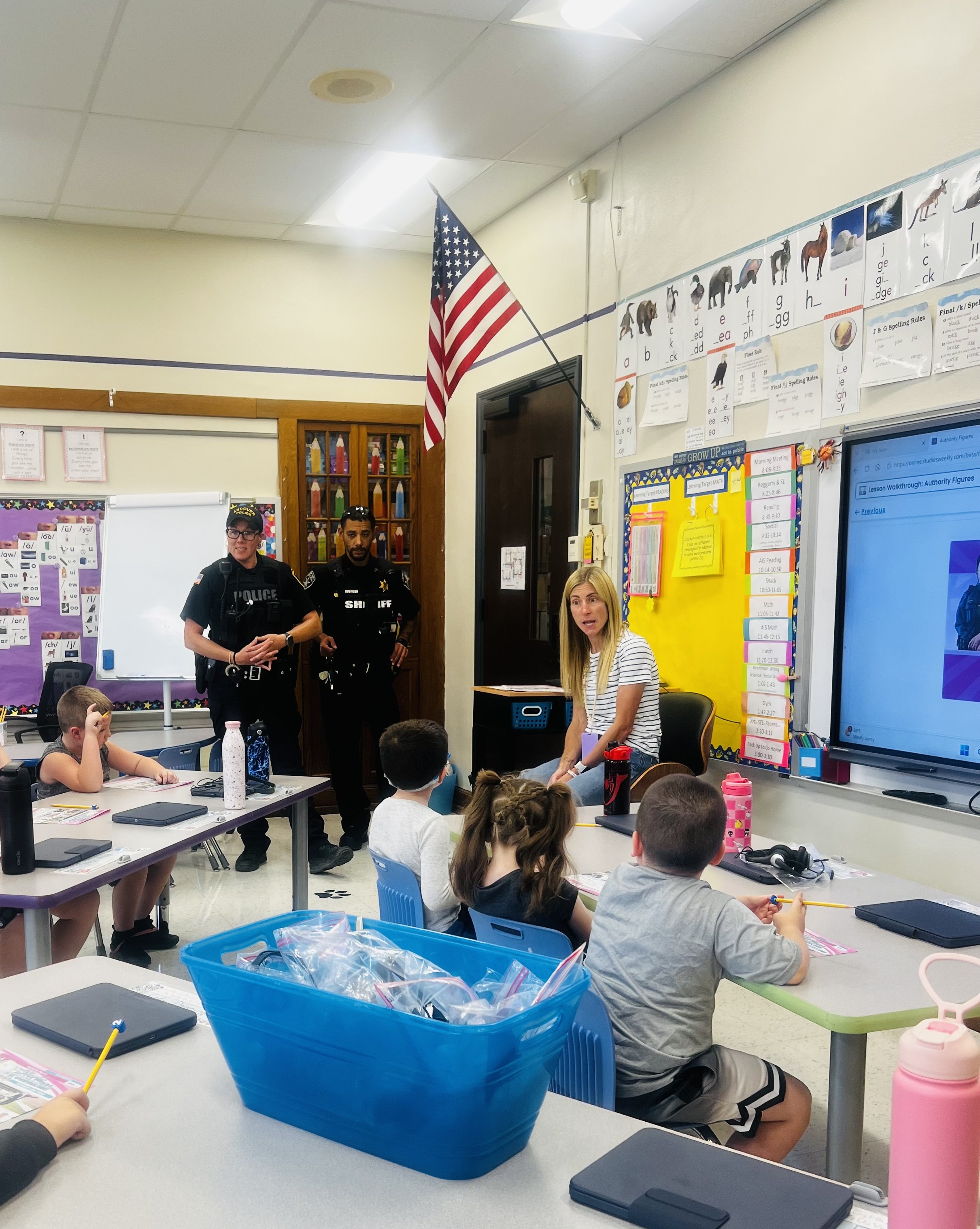 A teacher and law enforcement officers speak to students seated at desks in a brightly lit classroom.