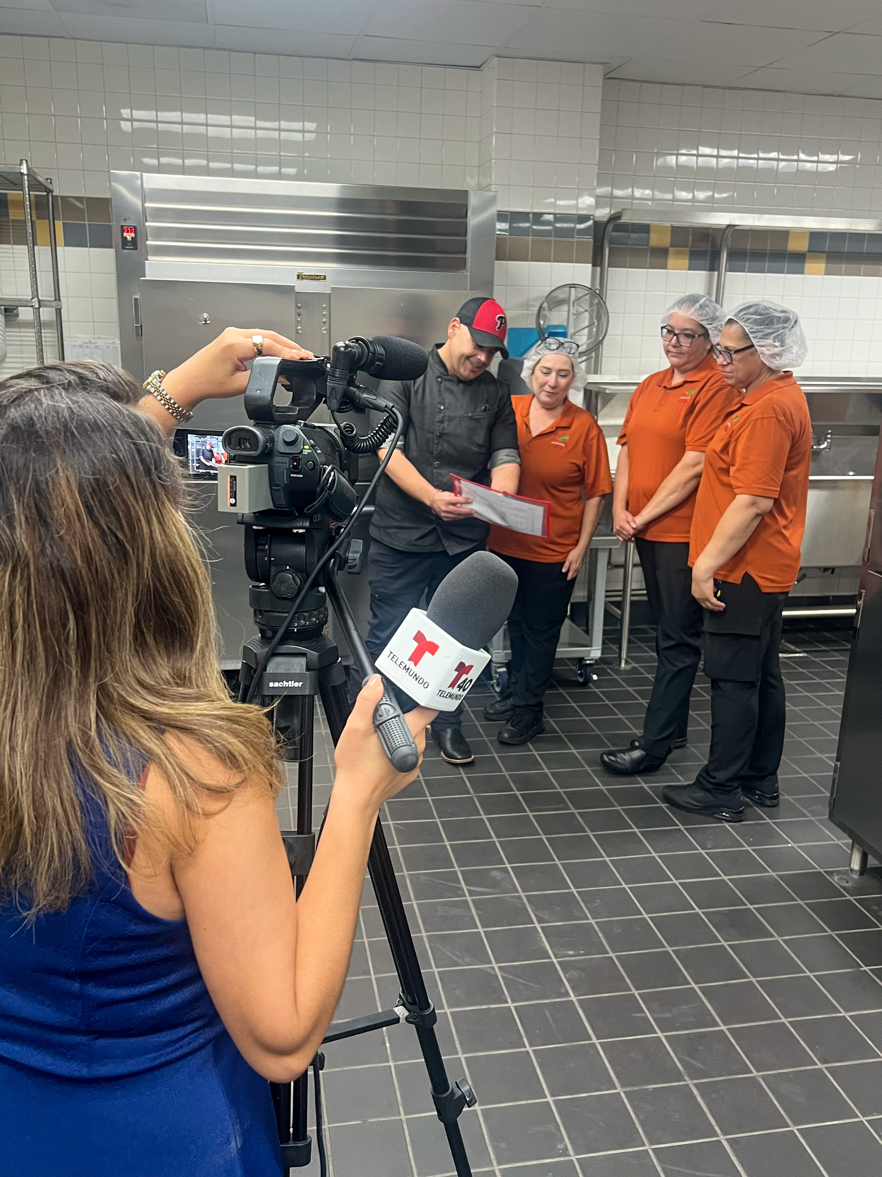 A news crew films a chef and three employees in a commercial kitchen.