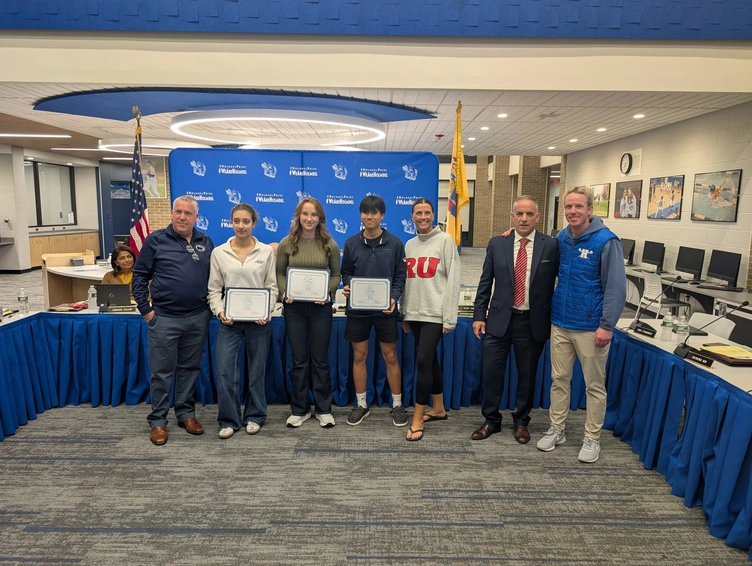 A group of adults and students stand together, holding certificates in a room with blue drapes and school banners.