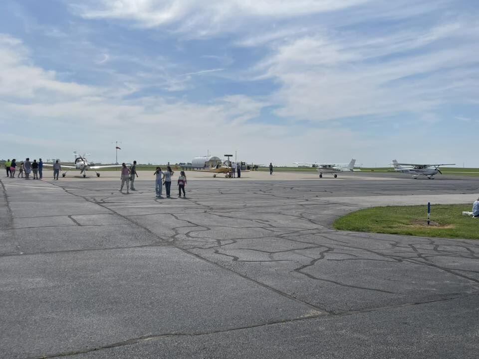 People gather on an airport tarmac, observing several small airplanes parked under a partly cloudy sky.