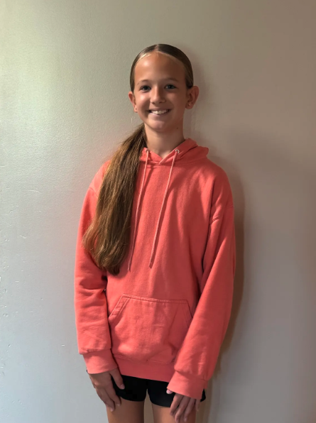 A smiling child with long brown hair stands against a neutral wall, wearing a peach-colored hoodie and black shorts.