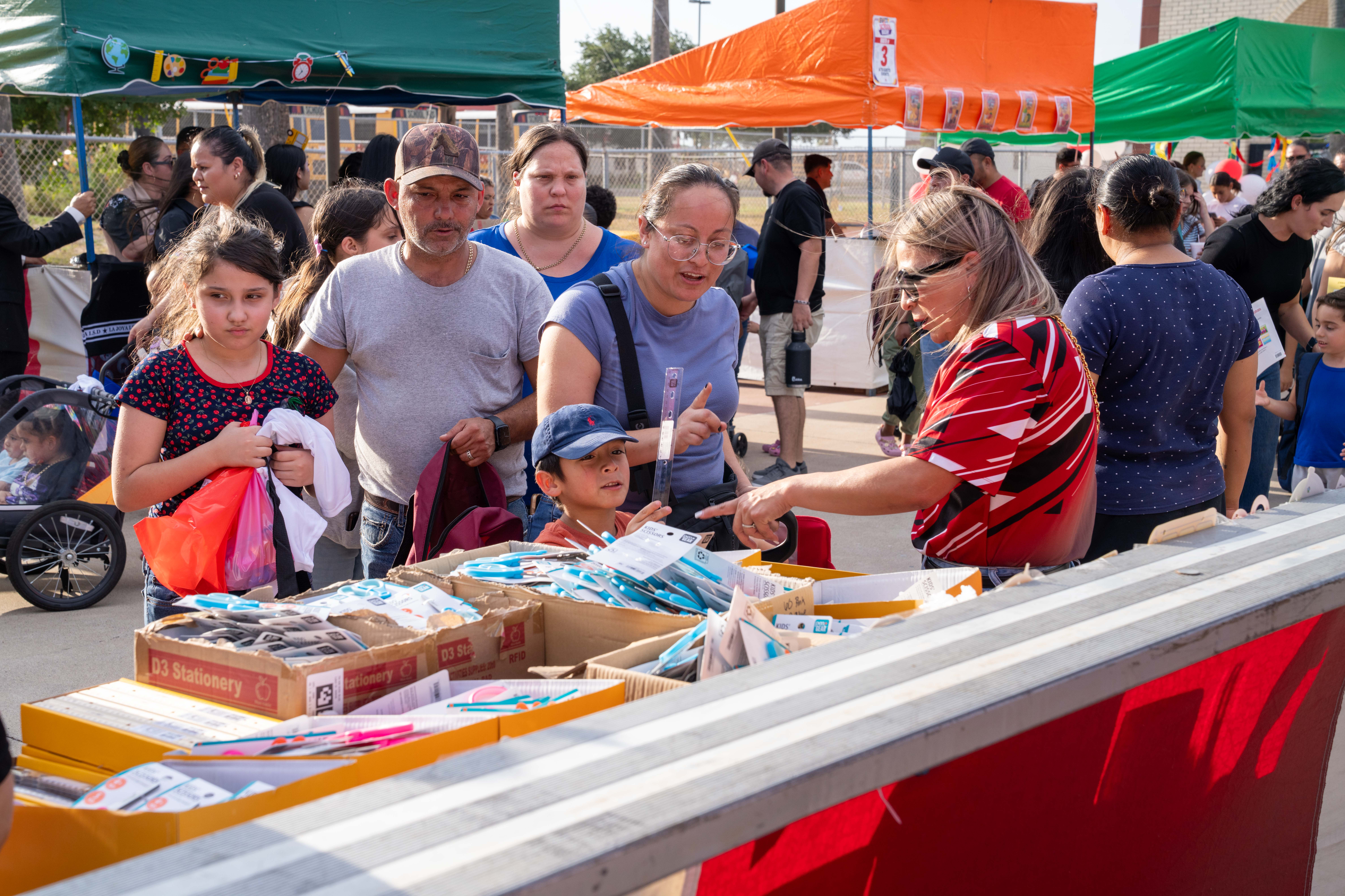 A diverse group of people gathers outdoors around tables filled with boxes of school supplies.