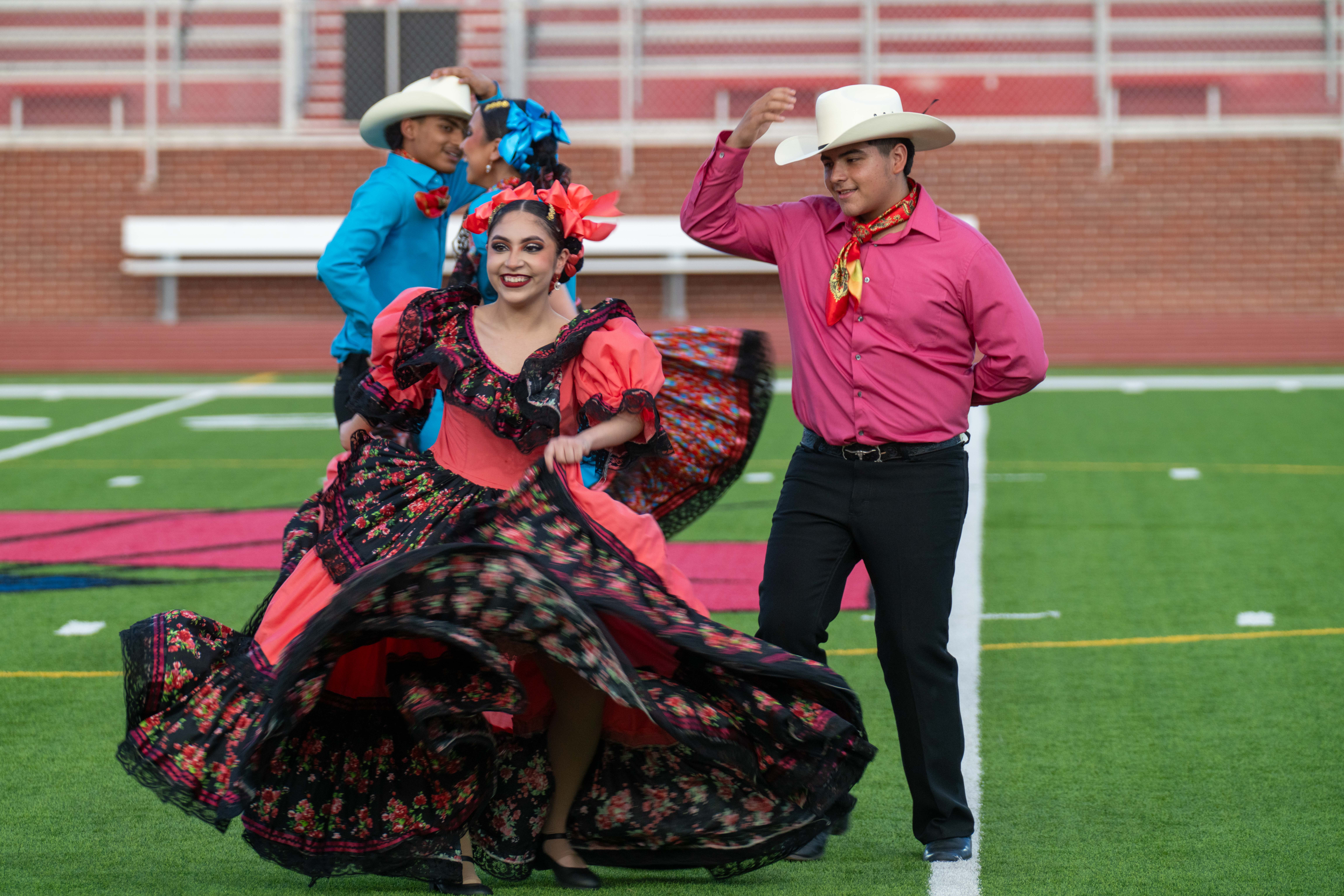 Dancers in vibrant traditional attire perform on a green athletic field.