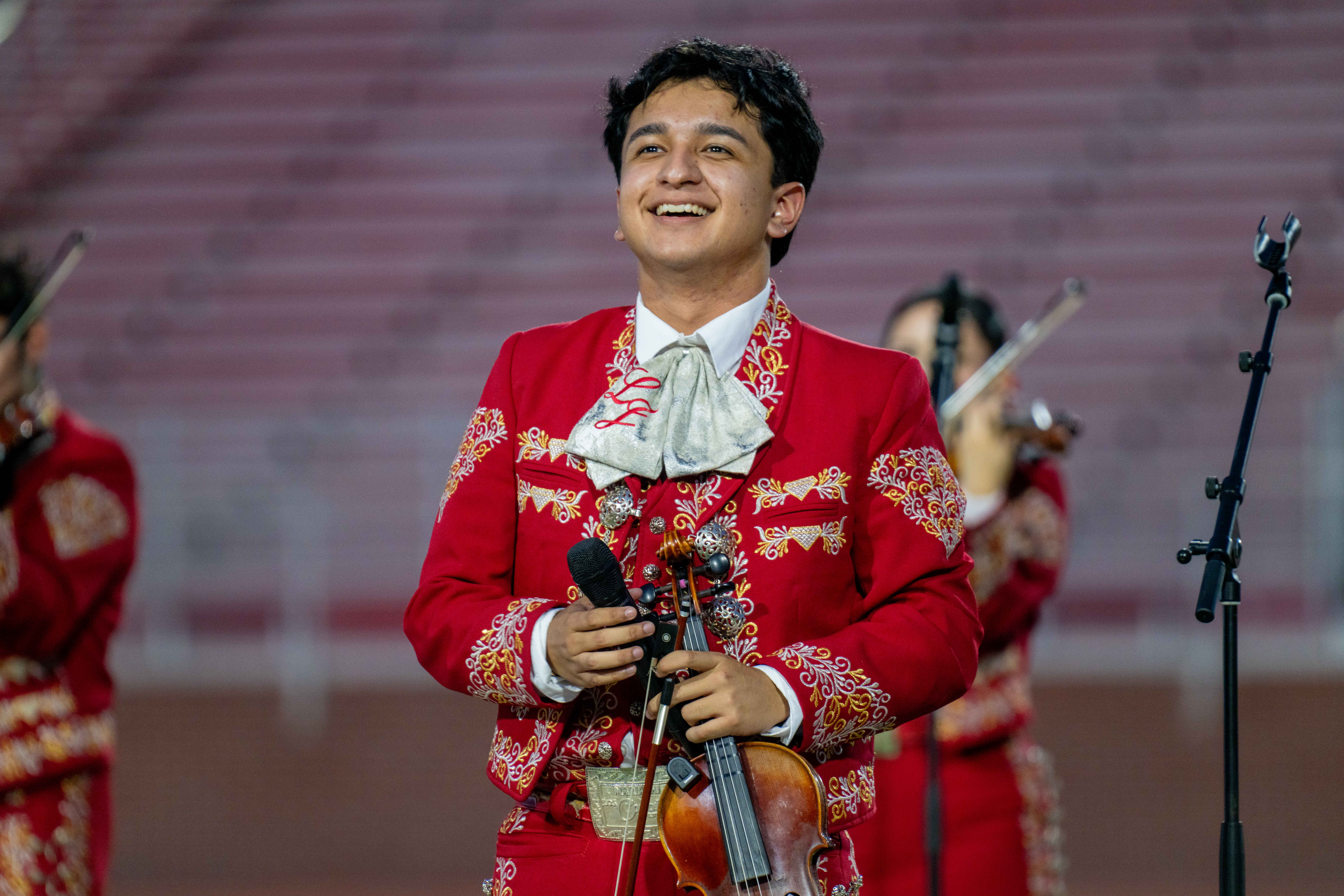 A smiling young musician in a red mariachi suit holds a violin and microphone outdoors.