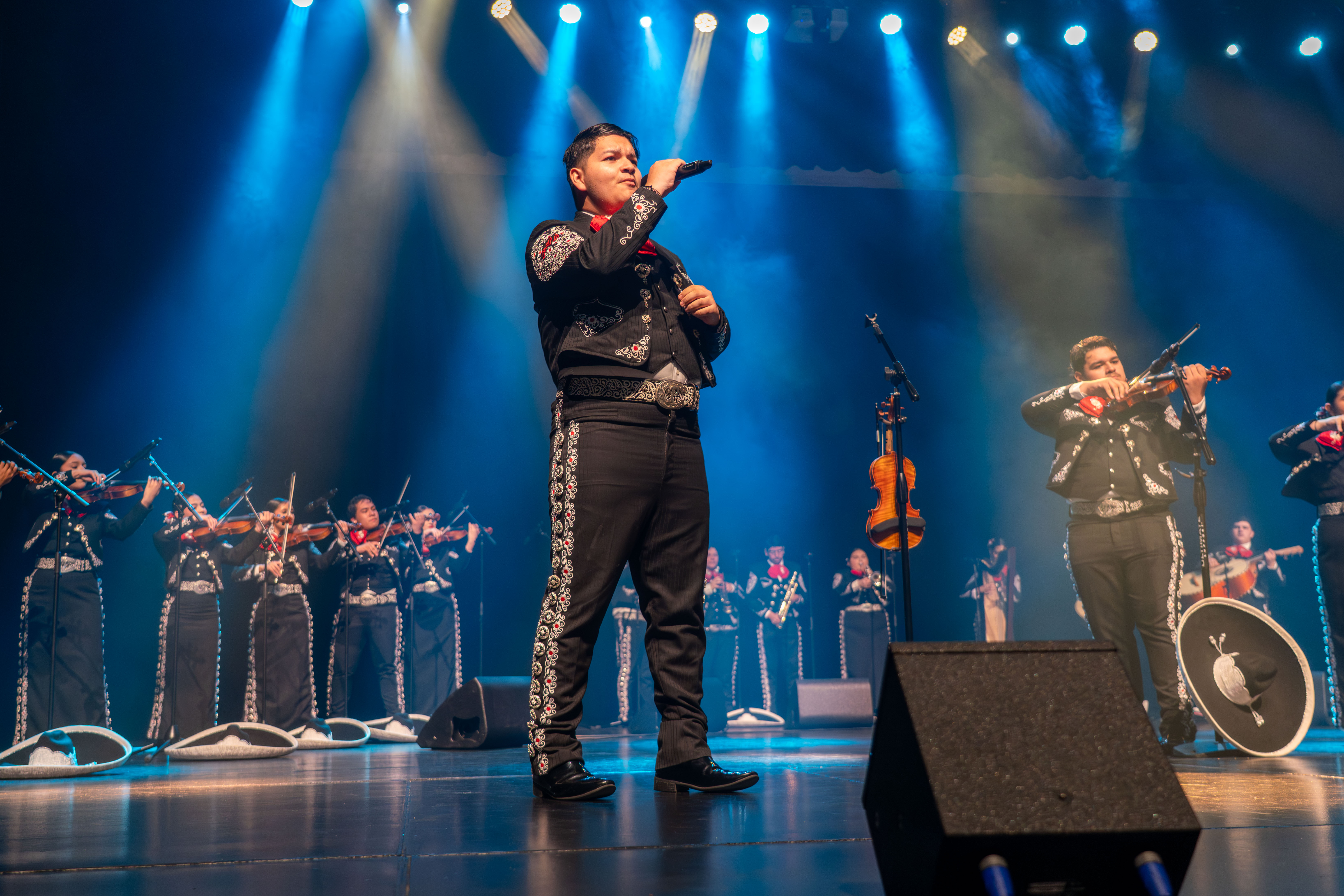 A mariachi singer holds a microphone on stage, with musicians playing violins and trumpets behind him.