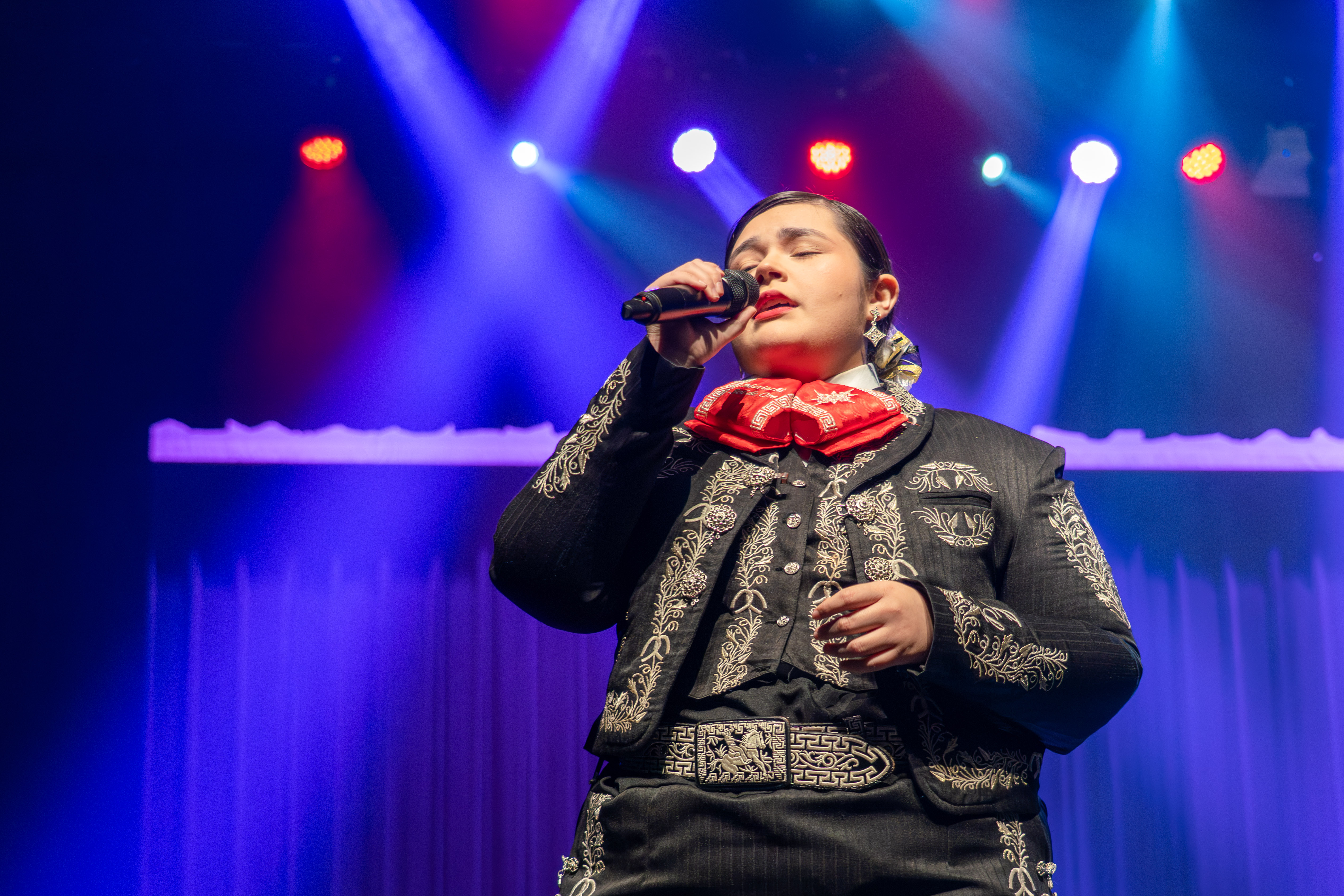 A performer in a traditional charro suit sings into a microphone on a stage with blue and red lights.