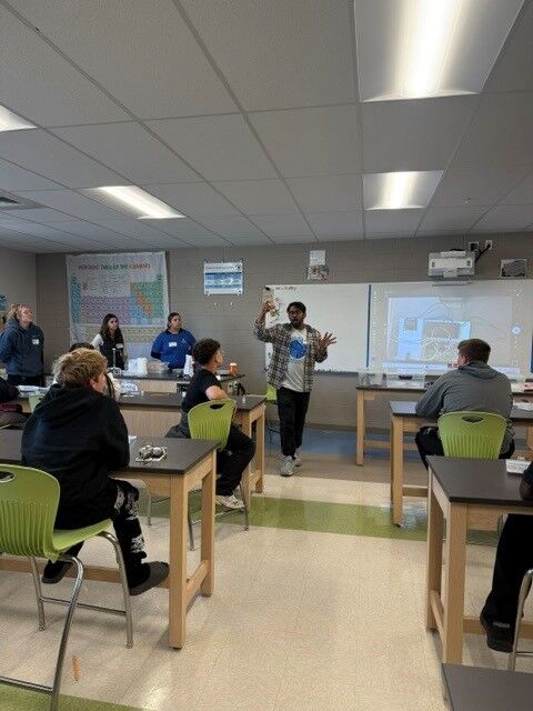 A teacher stands in front of a high school science classroom, gesturing while lecturing students seated at lab tables.