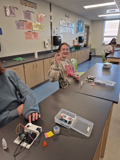 A smiling student sits at a science lab table, holding up a small component with electronic parts and a STEM kit nearby.