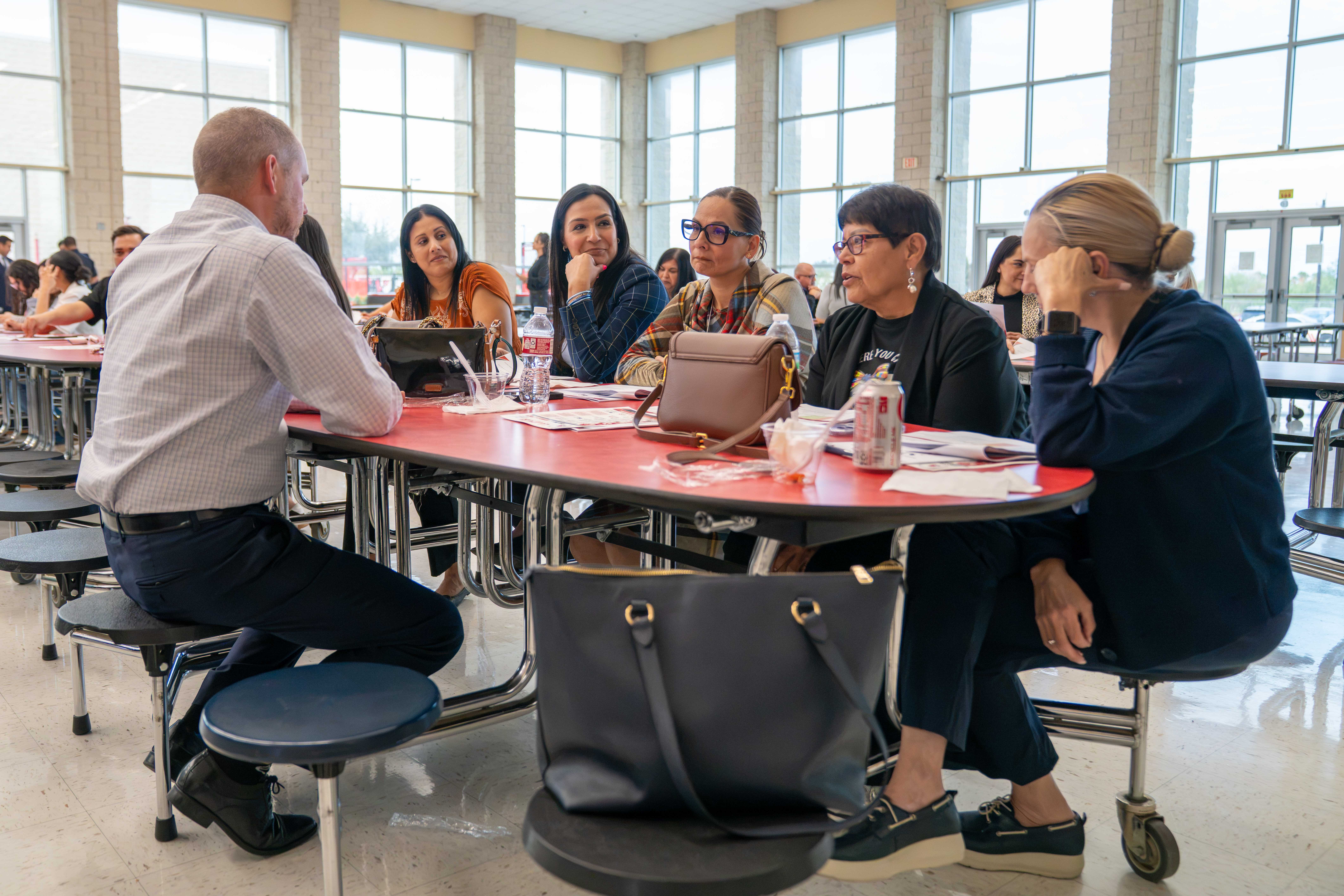 A group of people sit around a red cafeteria table, listening attentively in a bright, sunlit room.