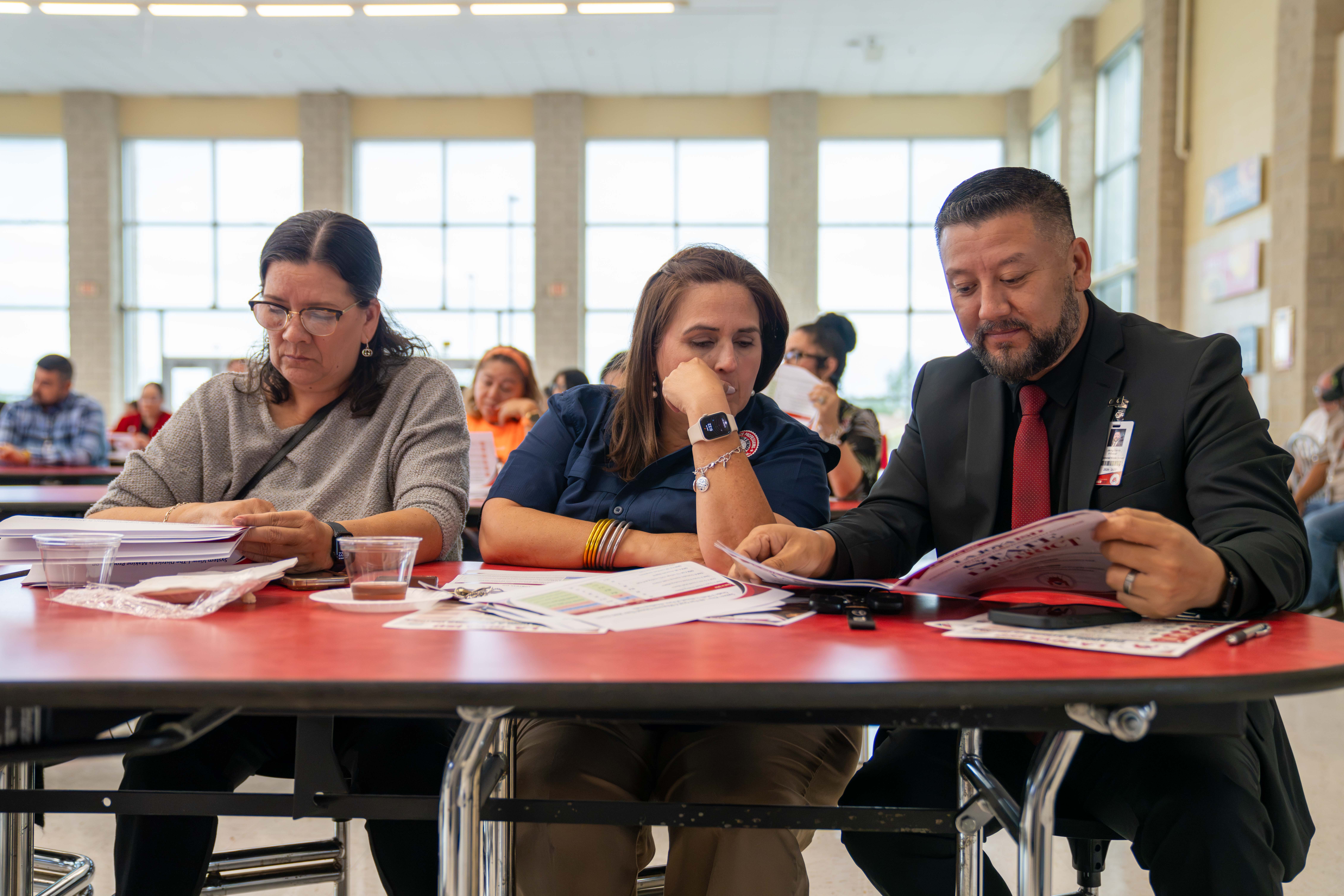Three people sit at a red table indoors, reviewing printed documents and listening intently.