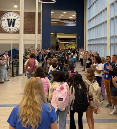 Students walk down a bright school hallway, greeted by staff applauding along the sides.