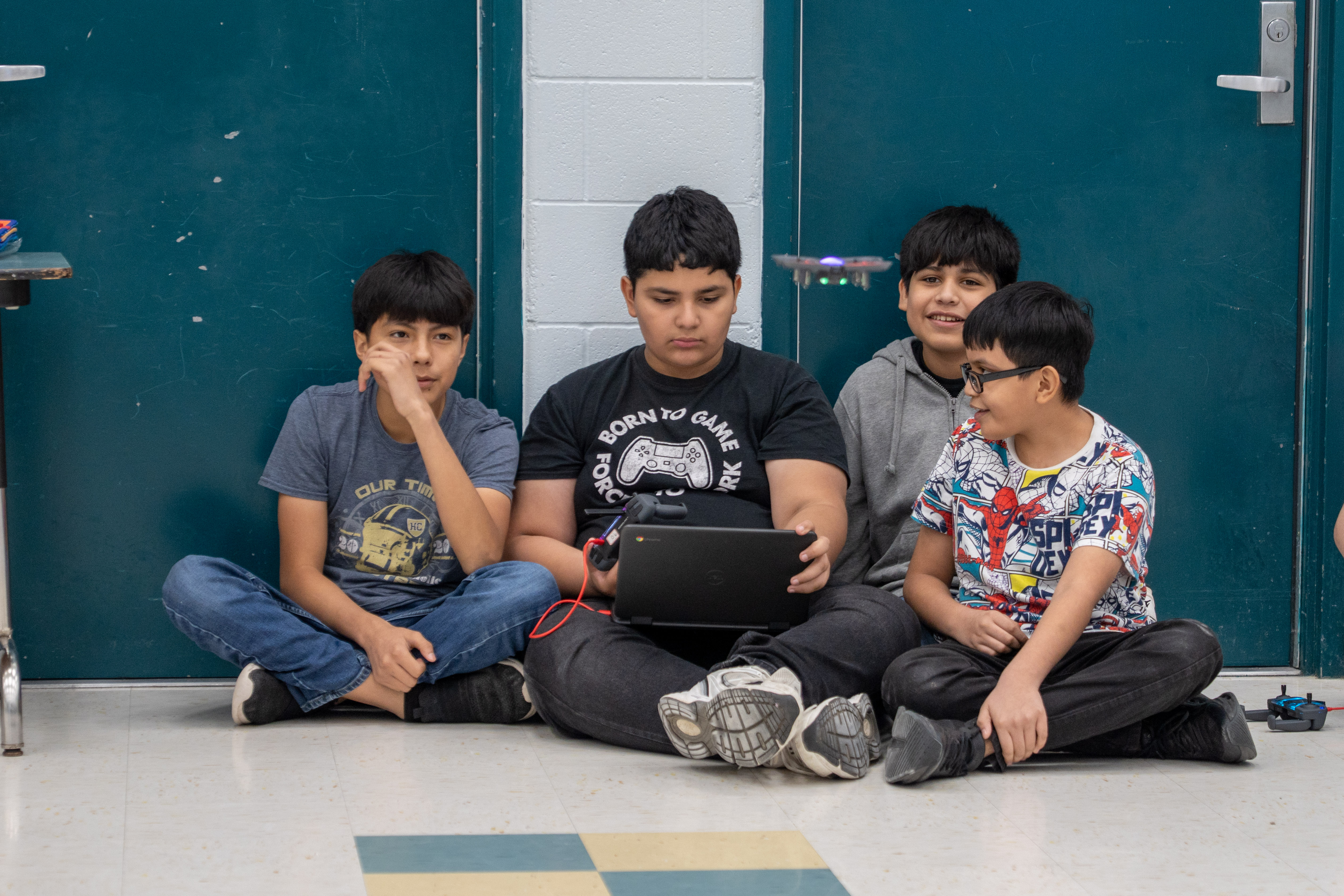 Four young boys sit on the floor, looking at a laptop and a drone in flight.