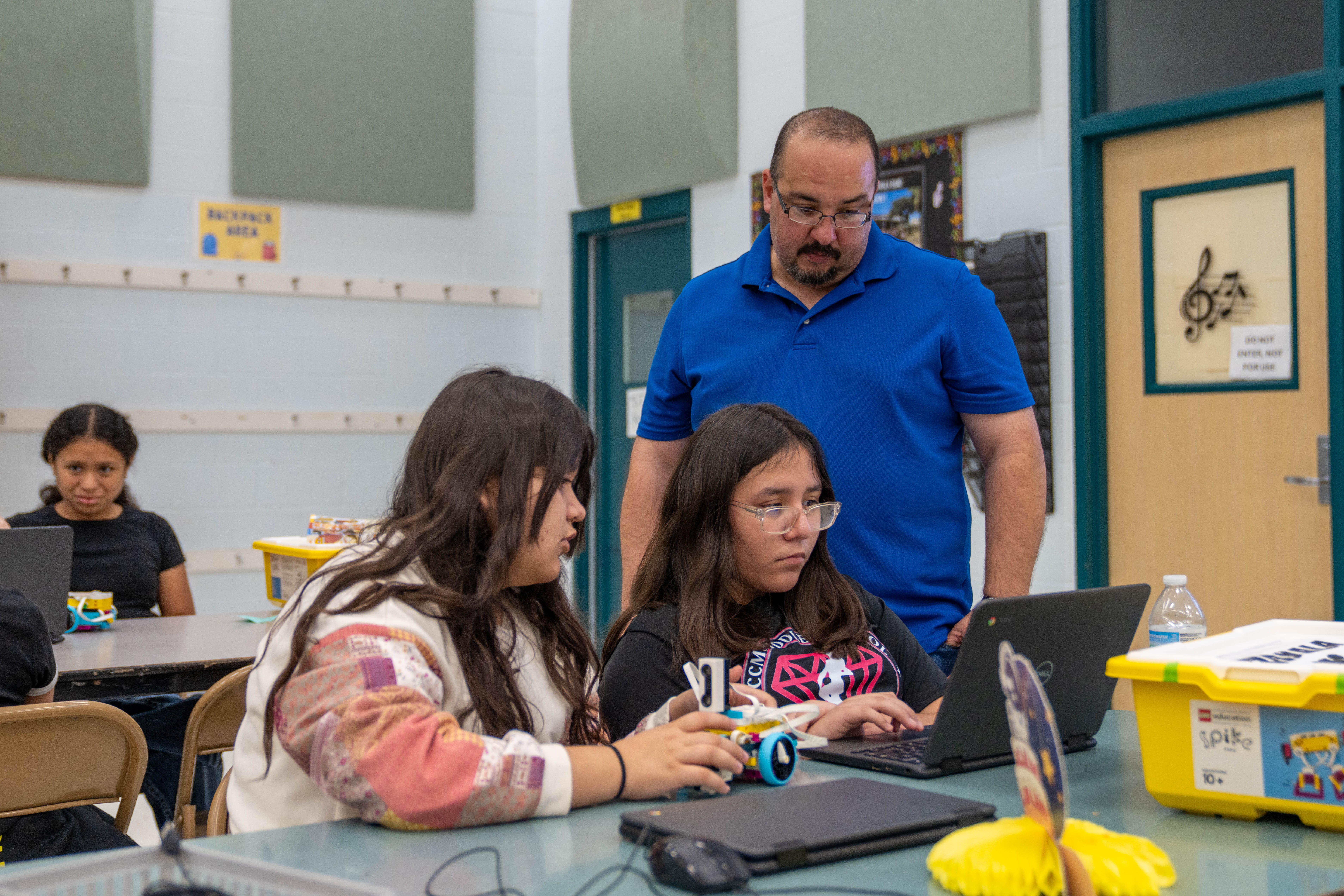 A teacher in a blue shirt observes two students working on laptops and a small robot.