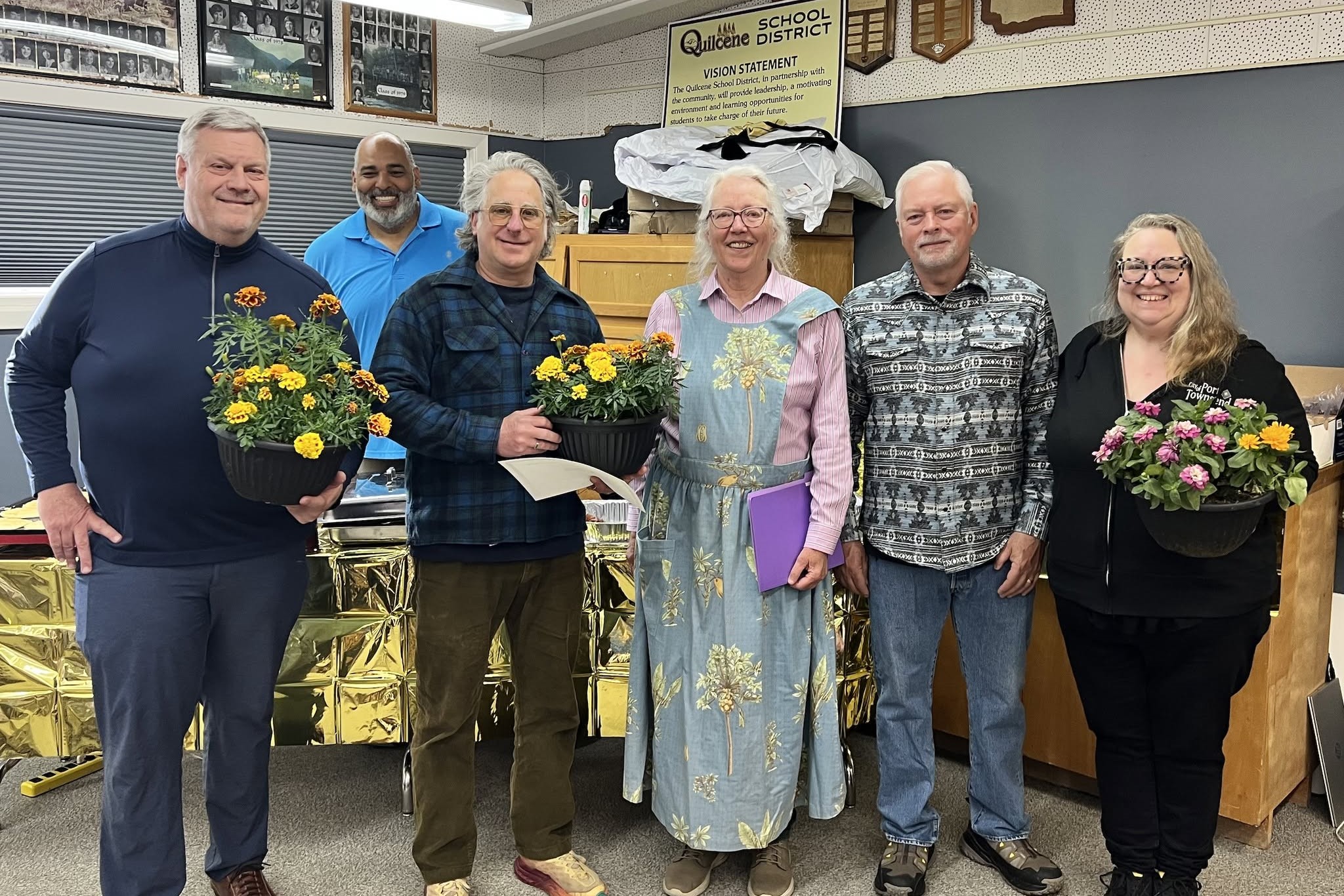 Six people stand together, holding potted flowers, in front of a gold-covered table and a school district vision statement.