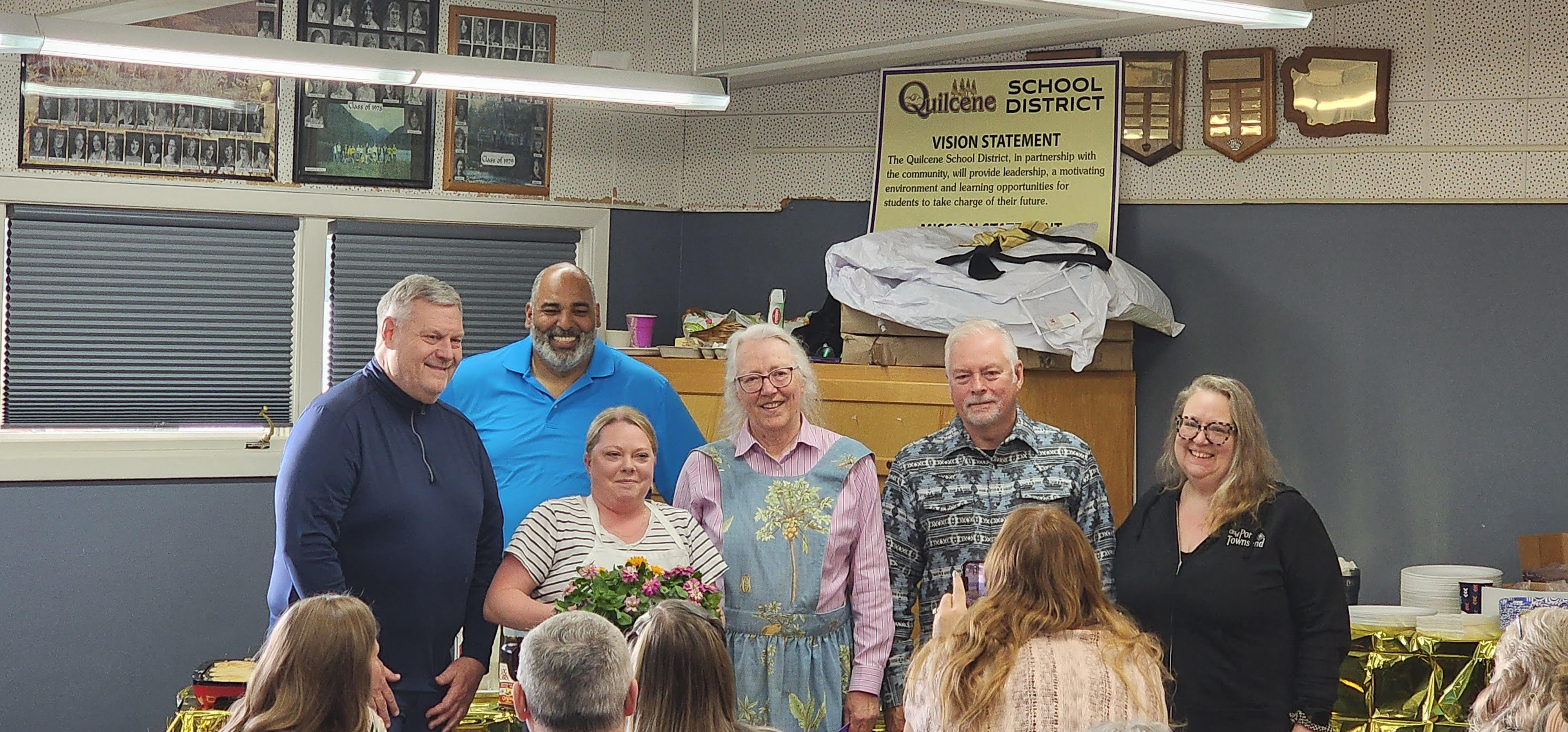 A group of people smile for a photo in front of a "Quilcene School District" vision statement poster.