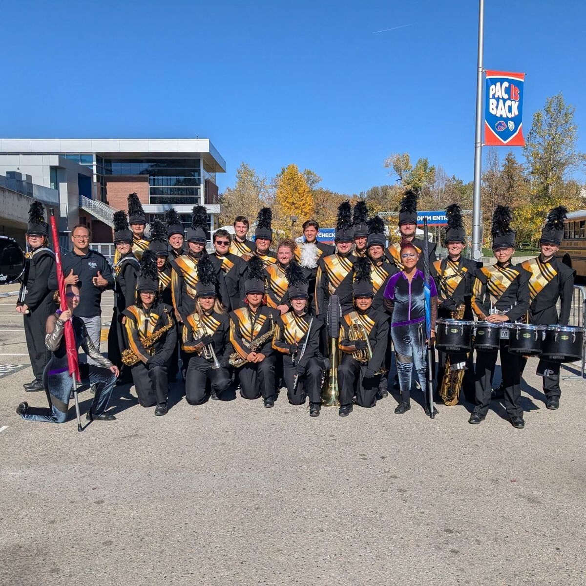A marching band in black and gold uniforms poses outdoors on a sunny day with instruments.