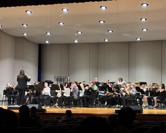 A conductor leads a school band performing on a stage with bright overhead lights.