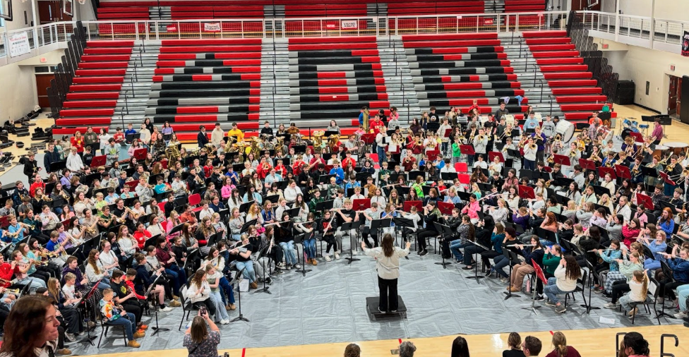 A large school band rehearses in a gymnasium with bleachers in the background.