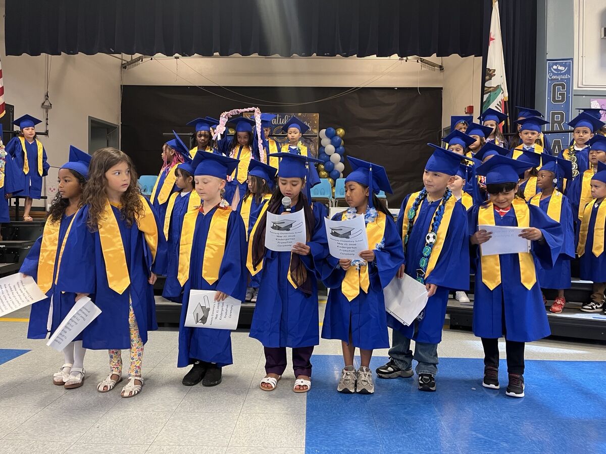 A group of young children in blue and gold graduation gowns and caps stand on a stage, holding certificates.