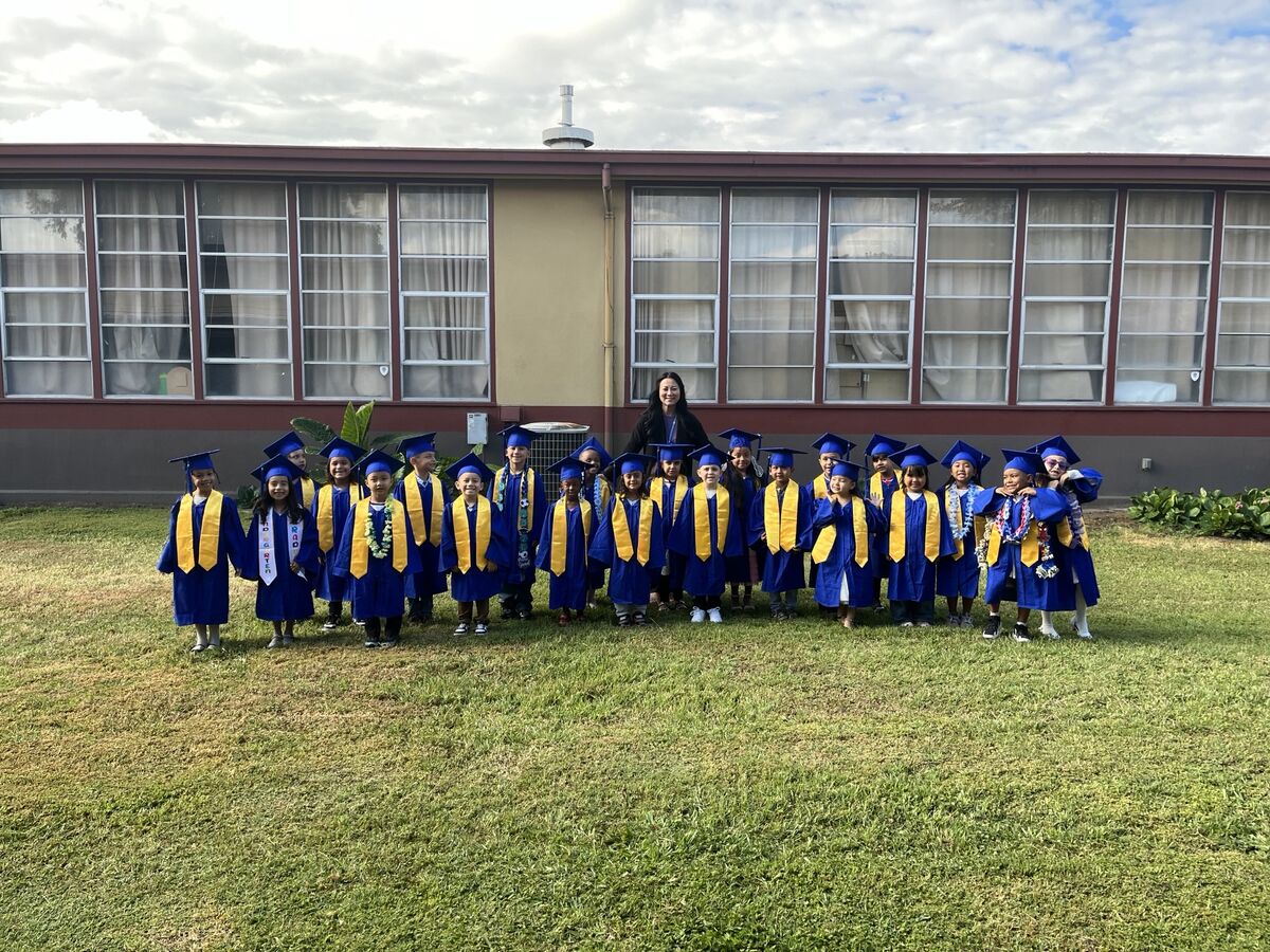 A group of young children in blue graduation gowns and caps stand on a grassy lawn in front of a school building.