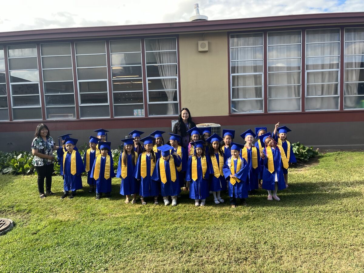 A group of young children in blue graduation gowns and caps stand on a grassy lawn in front of a school building.