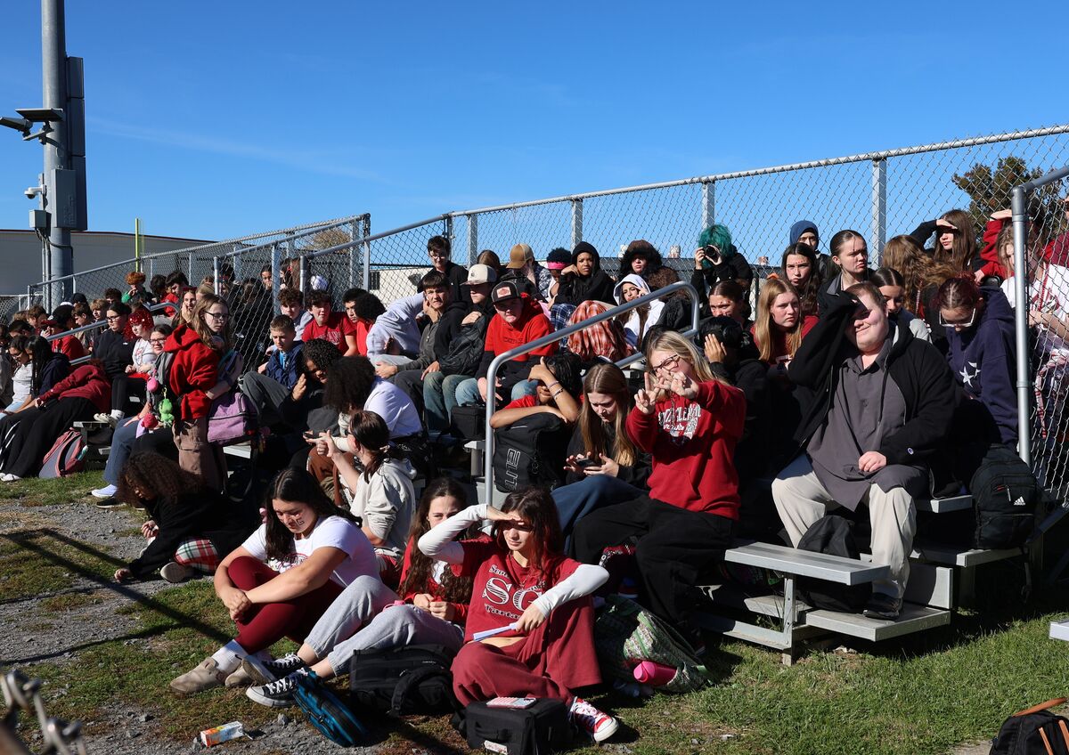 A large group of students sits and stands on metal bleachers outdoors, watching a game on a bright sunny day.