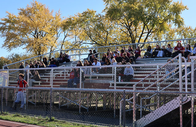 A large group of students fills outdoor metal bleachers on a sunny autumn day, with golden trees in the background.