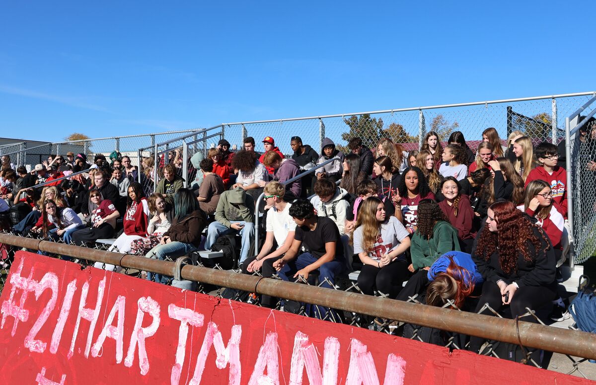 A large group of students sits in outdoor bleachers under a clear blue sky, watching an event.