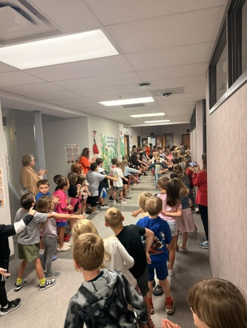 A long line of school children and staff line a brightly lit hallway, reaching out to high-five a student walking down the center.