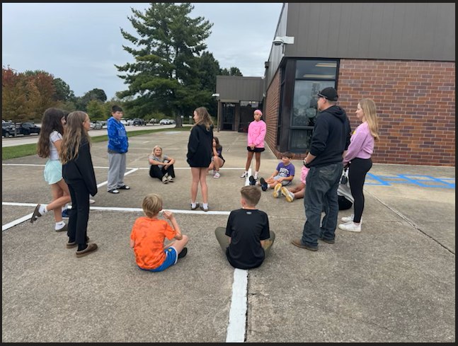 A group of children and adults gather outside on a paved courtyard near a brick building.