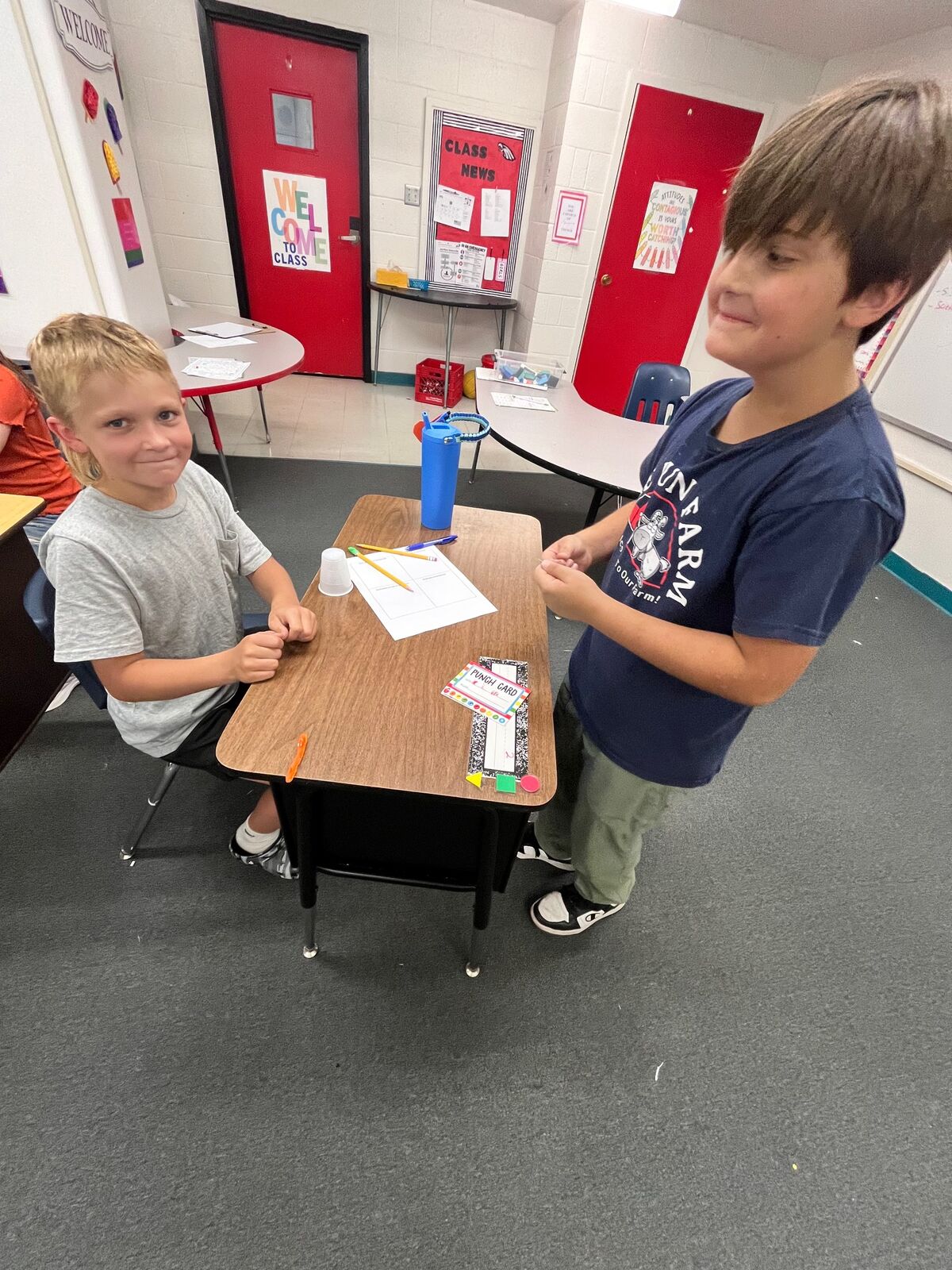 Two young boys are in a classroom, one standing and looking down at a desk, the other sitting and looking at the camera.