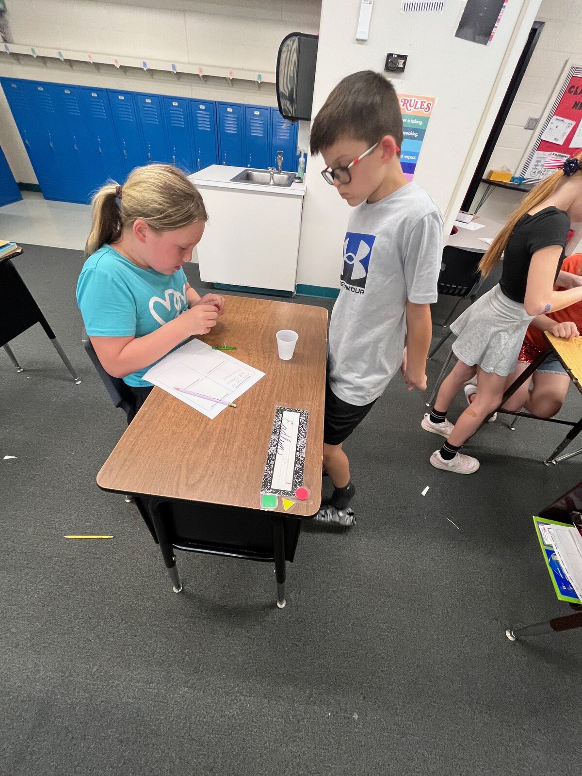 Students work at desks in a classroom with blue lockers in the background.