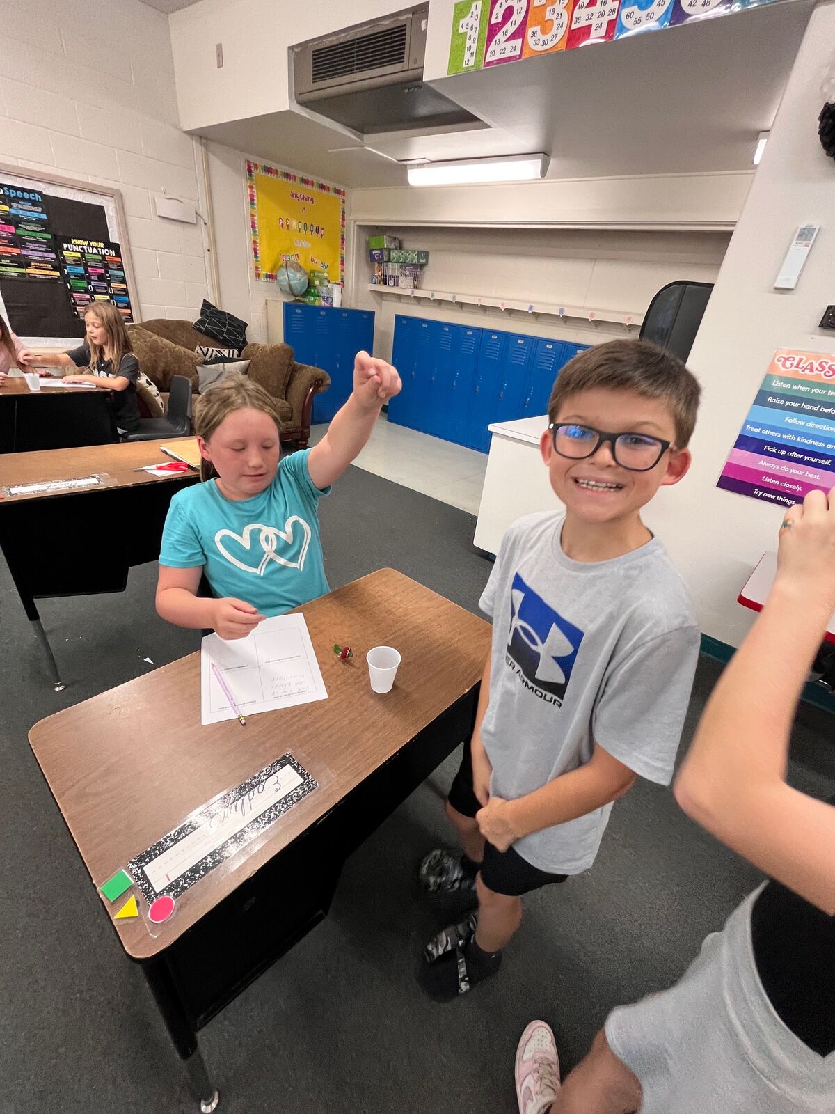 Two children are engaged in a classroom activity at their desks, with one raising their fist in excitement.