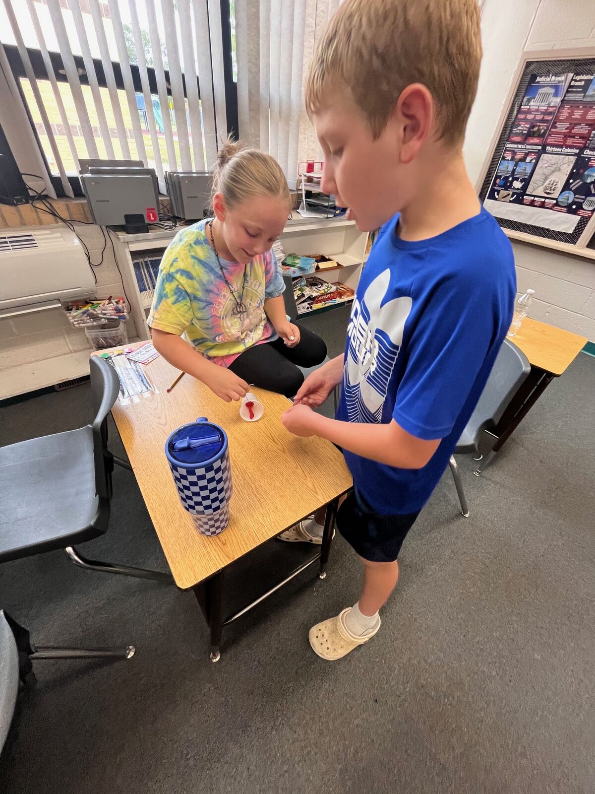 Two children are engaged in an art activity at a classroom desk, with one painting and the other observing.