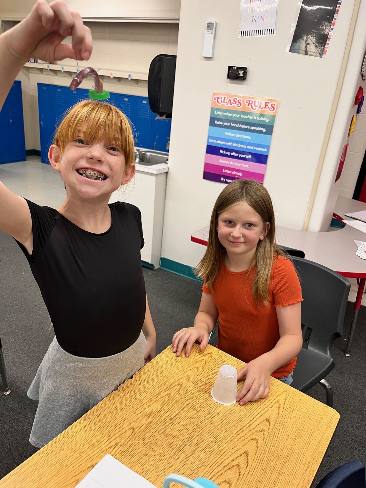 Two smiling children are in a classroom, with one holding up a small object.