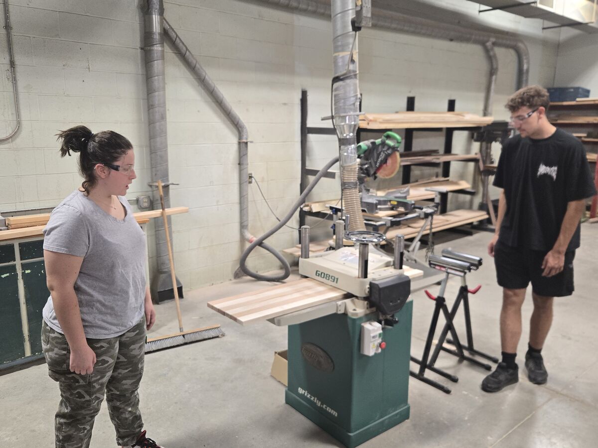 Two people stand in a workshop next to a Grizzly wood planer, with a piece of wood resting on its table.