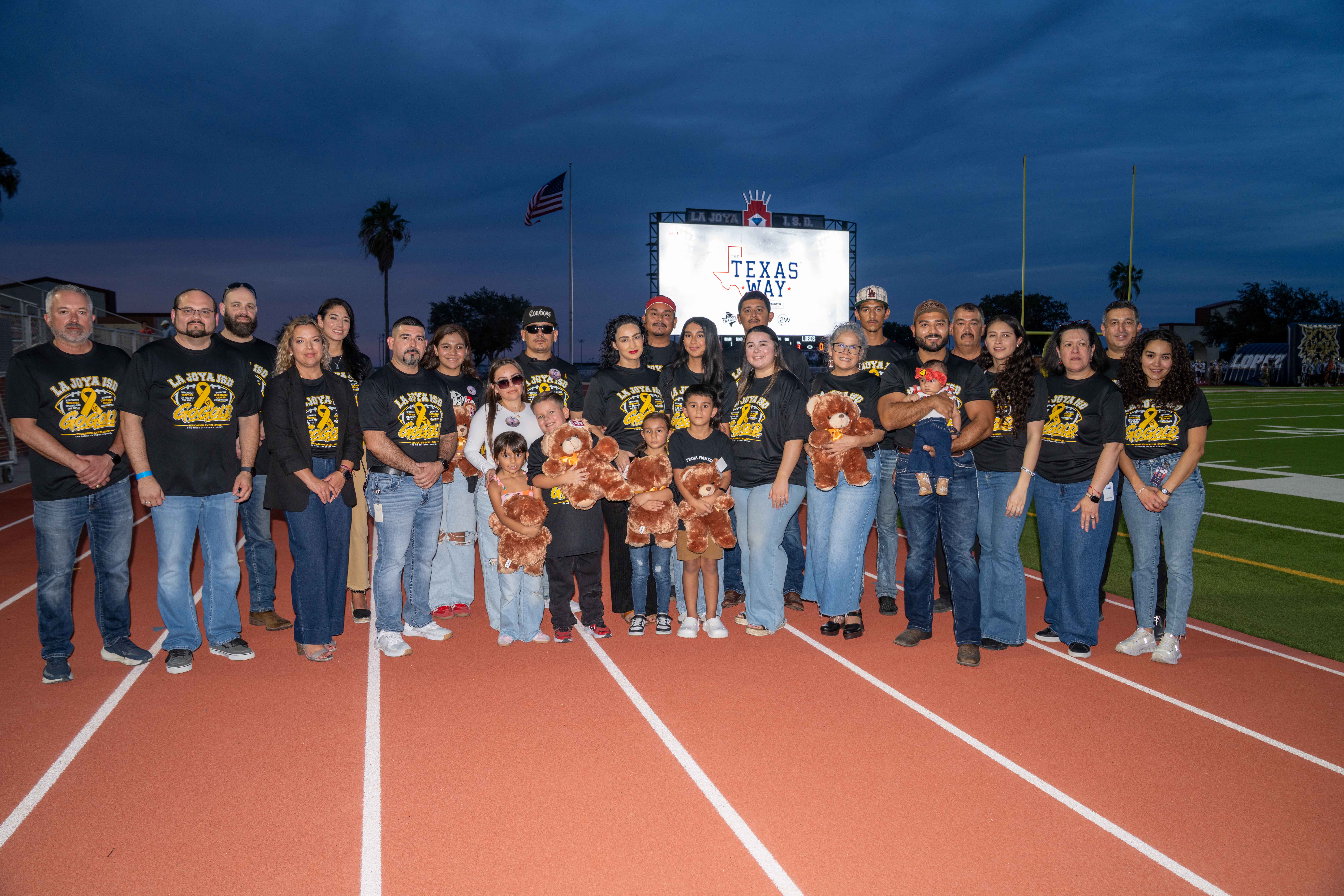 A large group of adults and children pose for a photo on an outdoor running track at dusk, with a lit scoreboard displaying "TEXAS WAY" in the background.