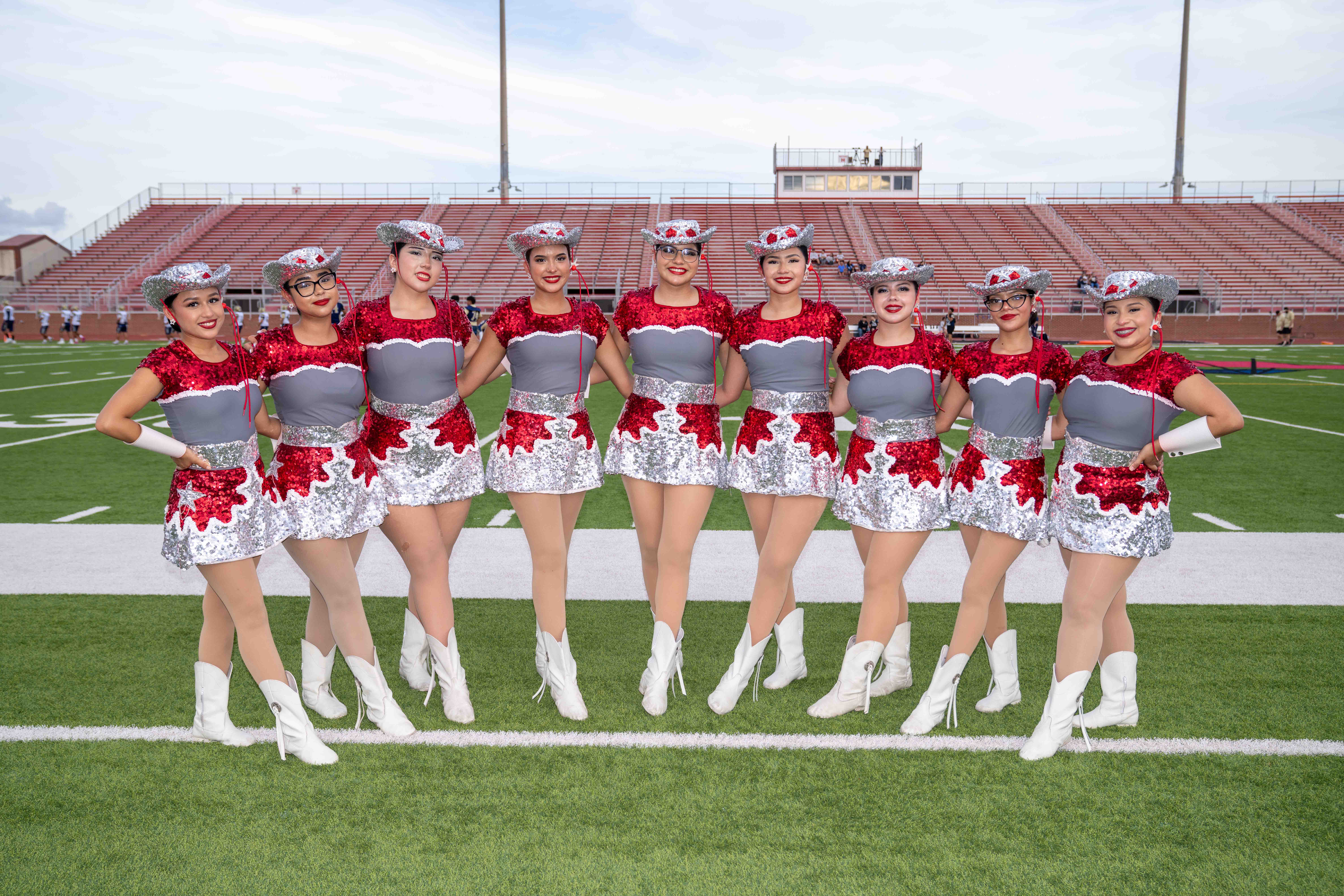 A line of nine smiling dance team members poses on a football field in matching red, grey, and silver sequined uniforms and cowboy hats.