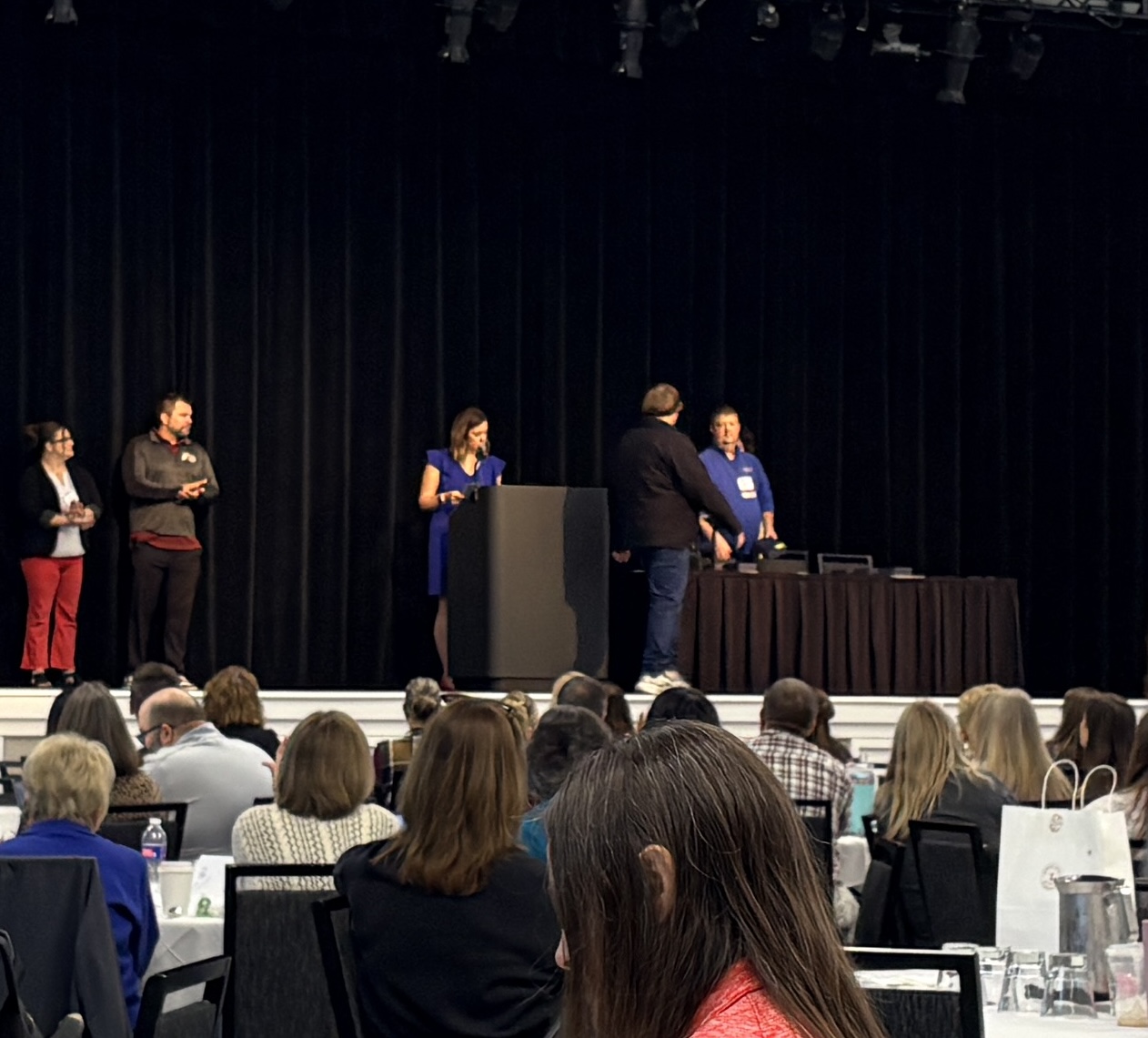 A speaker at a podium on stage presents to an audience seated at tables in a dimly lit auditorium.