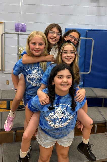 Five smiling girls in matching blue tie-dye shirts with 'Lancaster Wildcats' text are posing together.