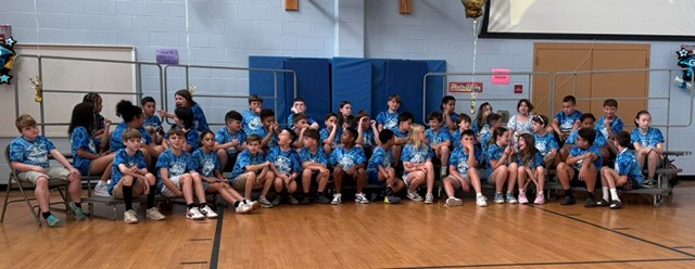 A group of children in matching blue tie-dye shirts sit on bleachers in a gymnasium.
