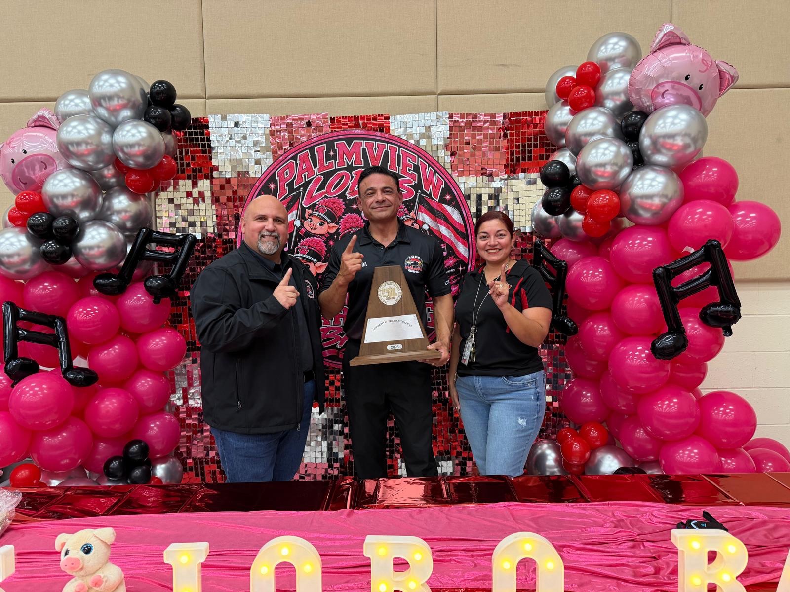 Three adults pose proudly, holding a trophy in front of a backdrop decorated with pink and silver balloons and musical note cutouts.