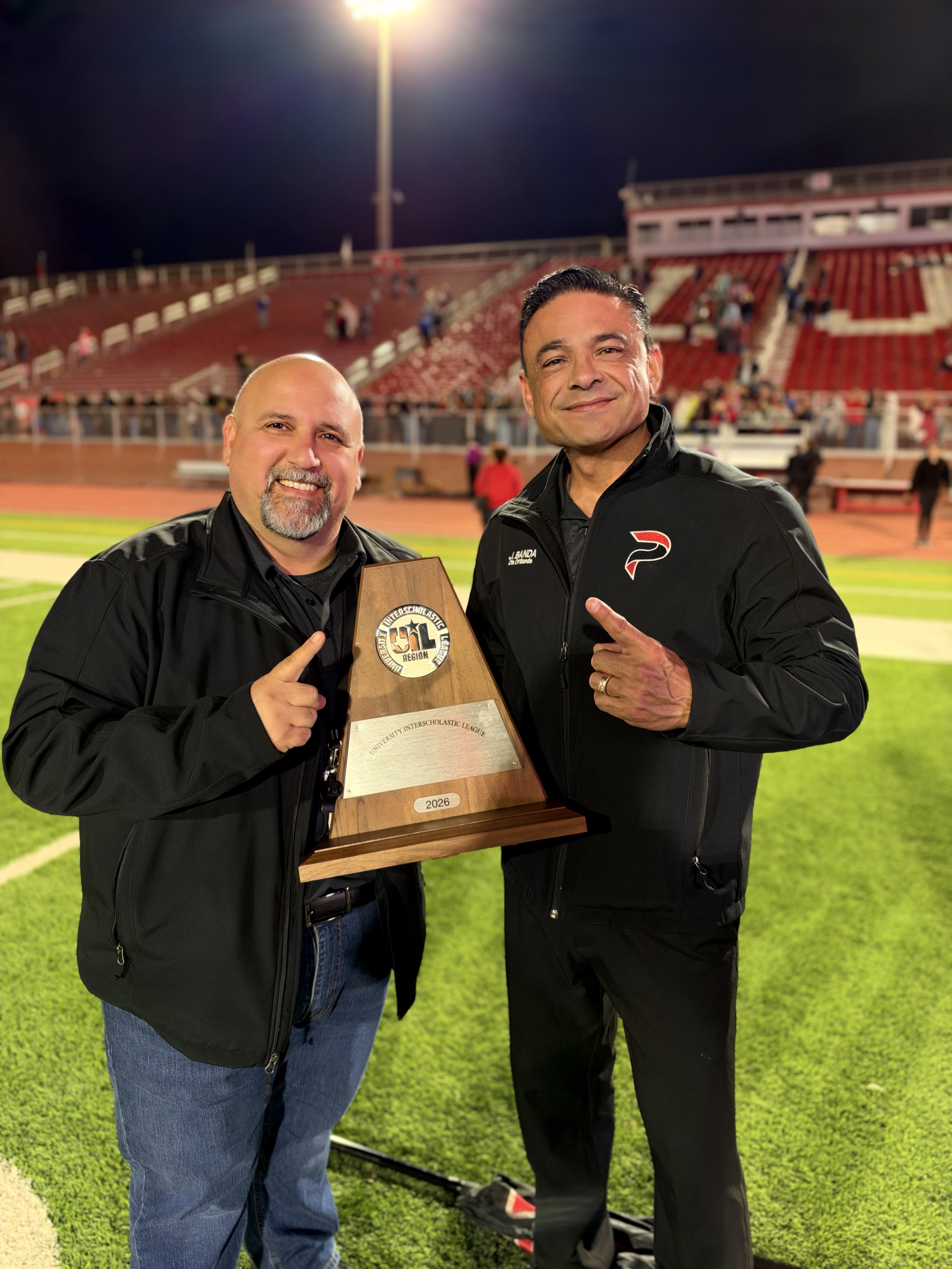 Two smiling men hold a University Interscholastic League 2026 region trophy on a football field at night.
